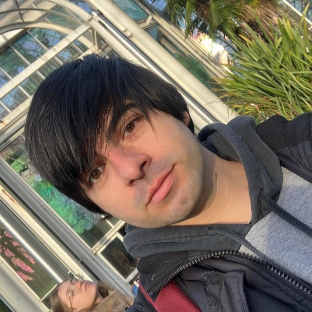 Tomi mirando a cámara, con un invernadero de vidrio y plantas detrás.